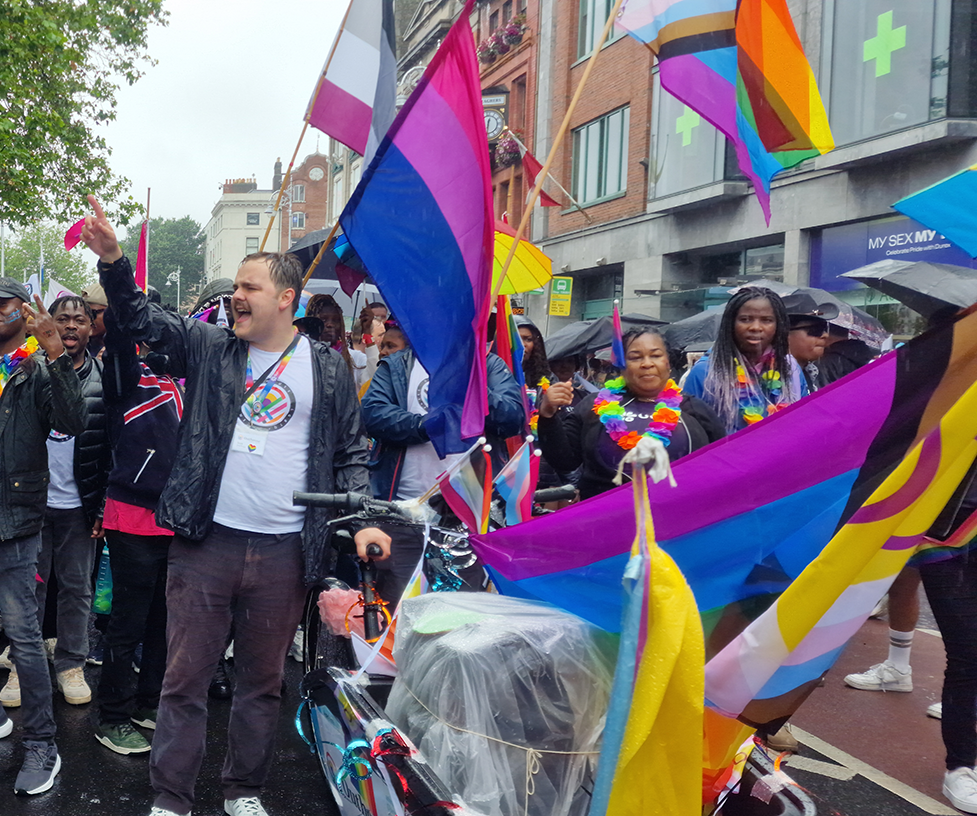 Person giving a speech at a Pride parade, standing in front of an Outhouse banner with Pride flags around them.